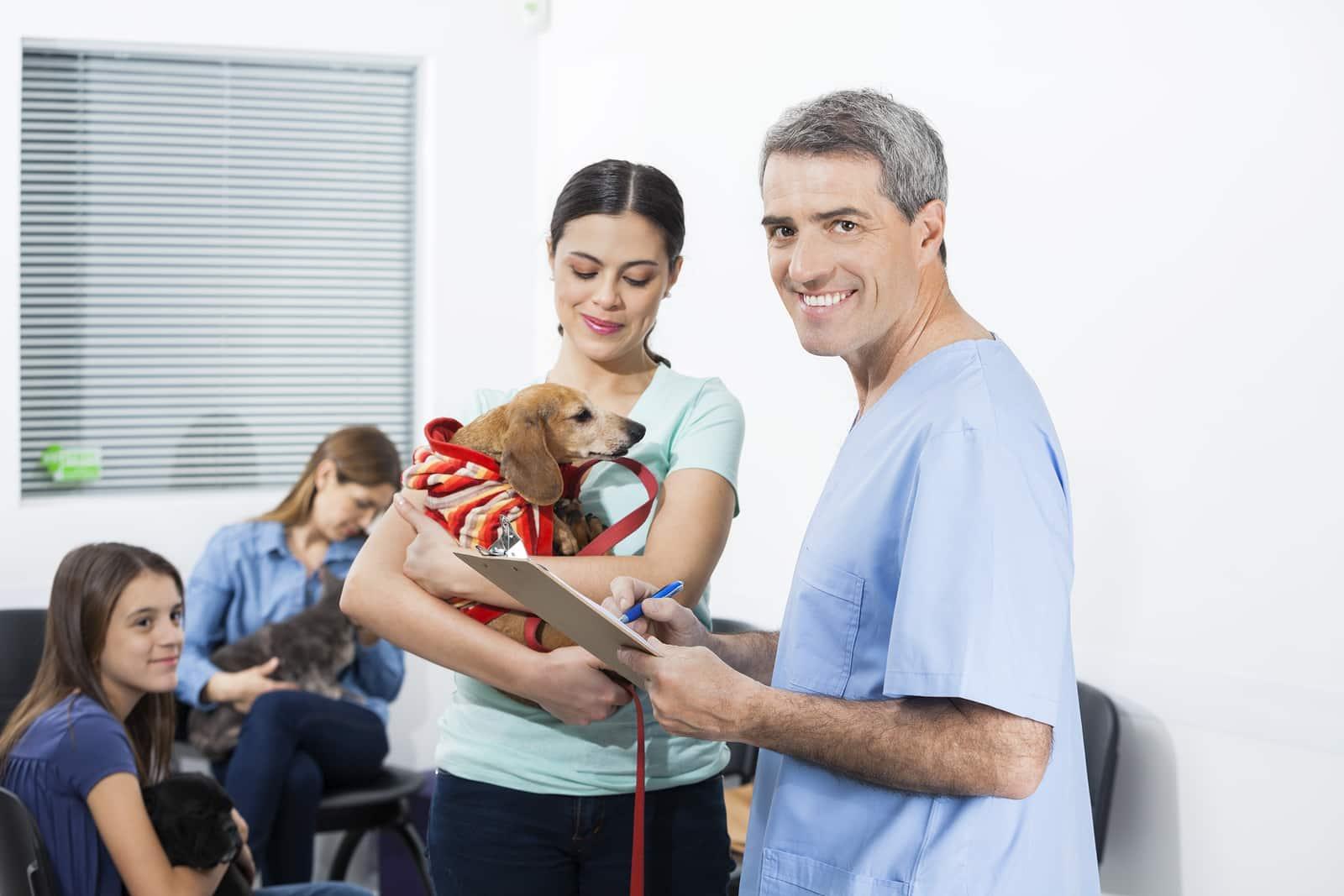 Male nurse in blue scrubs writes on clipboard while woman holds dachshund in waiting room.